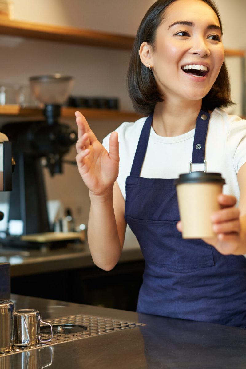 Portrait of smiling asian girl barista, giving out order in cafe, inviting guest to pick up takeaway order near counter, holding takeaway cup of coffee.