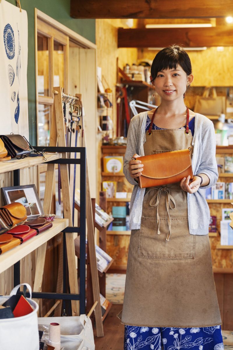 Japanese woman wearing apron standing in a leather shop, holding tan leather clutch bag, smiling at camera.